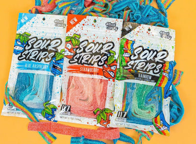 Three packets of sour strips candy are placed on a yellow surface with some unwrapped strips scattered around them. The flavors shown are Blue Raspberry, Strawberry, and Rainbow. Each packet has vibrant, colorful packaging and the strips are visibly sugar-coated.