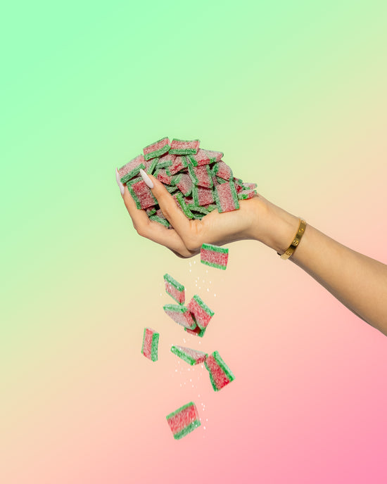 A hand with long nails and a gold bracelet drops colorful Sour Strips Bites-Four Pack red and green candies from Sour Strips against a pastel gradient background.