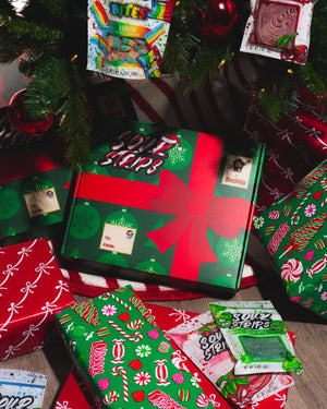 A festive scene features wrapped Christmas presents and candy under a decorated tree. Among them, the SourStrips Holiday Gift Box-Fourteen Pack stands out, adorned with a red bow and limited edition stickers for a special touch.