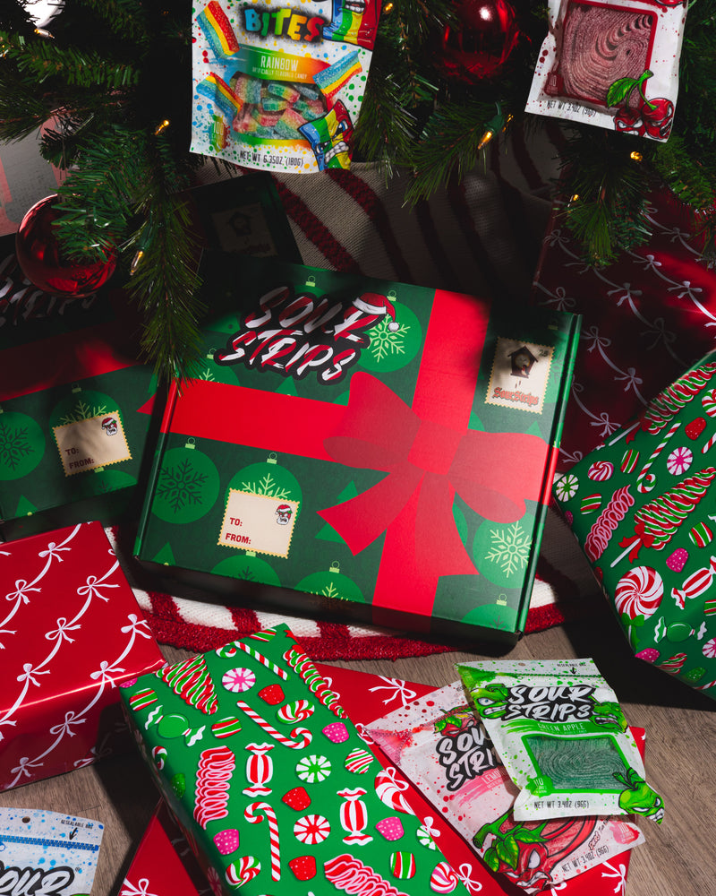 A festive scene features wrapped Christmas presents and candy under a decorated tree. Among them, the SourStrips Holiday Gift Box-Fourteen Pack stands out, adorned with a red bow and limited edition stickers for a special touch.