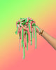 A hand displays a bunch of SourStrips Appleberry-Six Pack sour candy ribbons, red and green striped, against a lime green to soft pink gradient background. The person wears a gold bracelet.