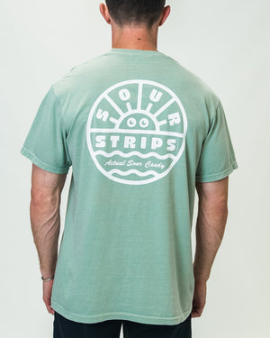 A person stands facing away, wearing the oversized box-fit Daylight Tee from SourStrips in light green. The unisex T-shirt features a white graphic design on the back with a smiling sun and the text "Sour Strips" and "Actual Sour Candy" below it. The person has short brown hair and is wearing dark pants.