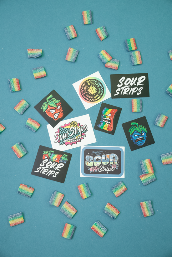 A variety of colorful, rainbow sour candy strips are scattered on a blue surface, surrounded by several kiss cut stickers from the SourStrips Sticker Pack. The stickers display various designs and text, including phrases such as "Sour Strips.