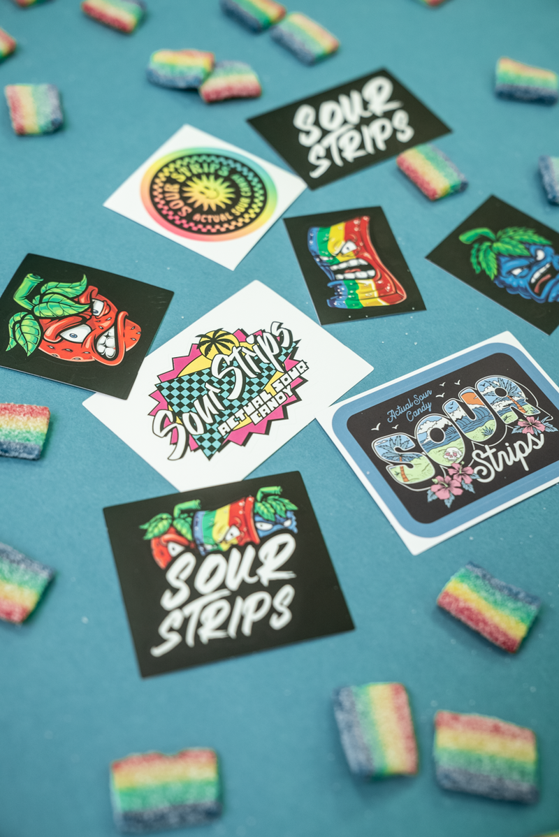 An assortment of colorful kiss cut stickers and candy pieces laid out on a blue surface. The stickers, part of the SourStrips Sticker Pack, feature vibrant designs with the text "SourStrips." The small, rectangular candy pieces are rainbow-colored.