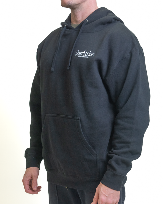 A person wearing a Retro Cherry Hoodie from SourStrips stands against a white background. This oversized black unisex design features "Sour Strips" on the chest, a front pocket, and drawstrings, showing the side profile from the waist up.