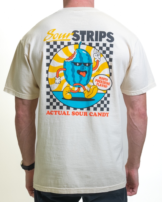 A person is wearing a Retro Watermelon Tee from SourStrips, an oversized beige unisex cotton T-shirt with a vibrant cartoon blue candy graphic on the back against a checkered background and text reading "Sour Strips Actual Sour Candy" and "Mouth Puckering Flavor.