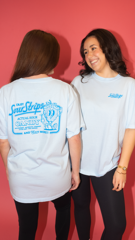 Two people wear light blue "Retro Blue Raspberry Tee" shirts by SourStrips against a pink background. The oversized box fit is showcased as one smiles facing forward, and the other displays the back design. Both pair their tees with black pants.