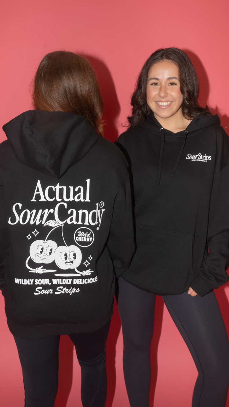 Two people against a pink background wear black Retro Cherry Hoodies from SourStrips. One faces the camera, smiling, while the other displays the back of their hoodie featuring a cartoon face and "Actual Sour Candy," combining unisex design with playful style.