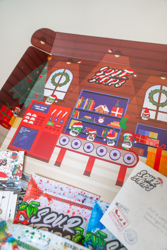 The SourStrips Holiday Gift Box-Fourteen Pack features a colorful illustrated lid with a festive factory, stockings, gifts, and wreaths. Sour Strips candy packs, branded paper, and exclusive limited-edition stickers are showcased in the foreground.