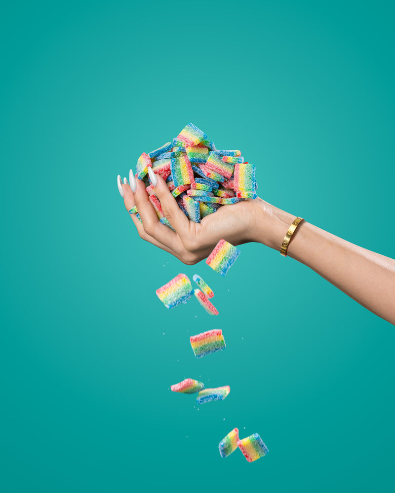 A hand with manicured nails, adorned with a gold bracelet, is holding a pile of colorful Sour Strips Bites-Four Pack by Sour Strips against a plain teal background. Some candy pieces are slipping through the fingers, falling down.