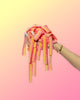 A hand with manicured nails holds a limited edition Lemonberry-Six Pack from SourStrips against a pink and yellow gradient background. These extremely sour, pink and yellow striped candies offer a tangy delight. The person is wearing a gold bracelet.