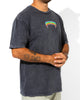 A person wears the SourStrips Sour Storm Tee—a unisex faded black cotton shirt with a colorful spiky logo on the left chest—paired with light pants and showing forearm tattoos. Their head is not visible.