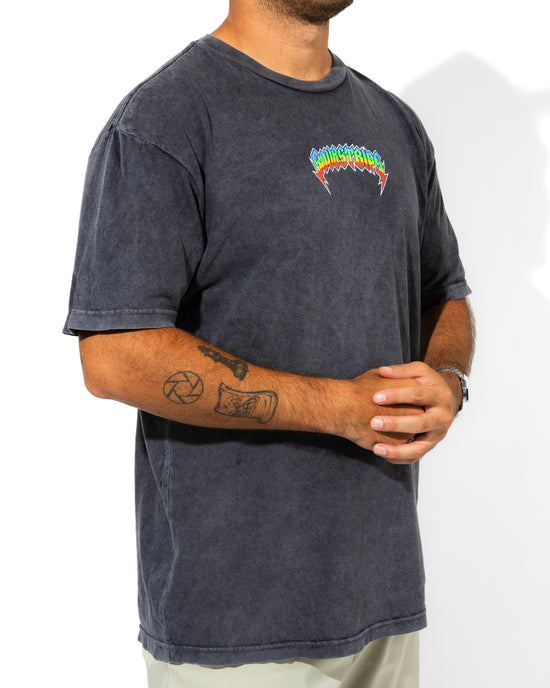 A person wears the SourStrips Sour Storm Tee—a unisex faded black cotton shirt with a colorful spiky logo on the left chest—paired with light pants and showing forearm tattoos. Their head is not visible.