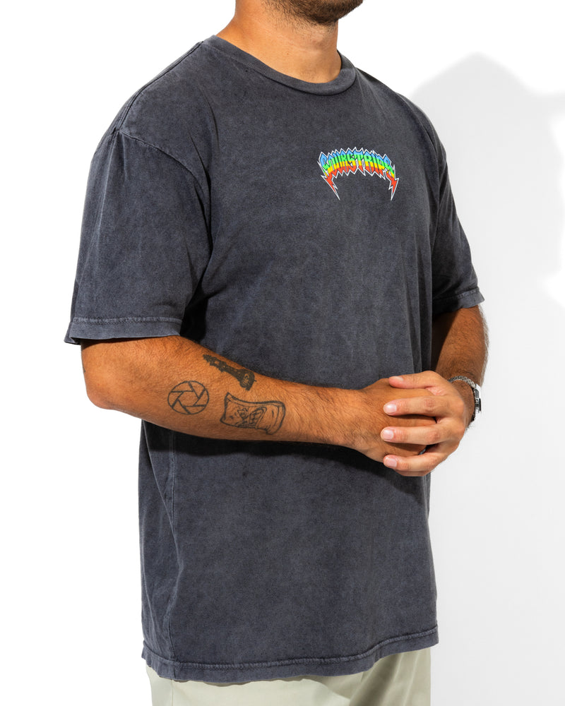 A person wears the SourStrips Sour Storm Tee—a unisex faded black cotton shirt with a colorful spiky logo on the left chest—paired with light pants and showing forearm tattoos. Their head is not visible.