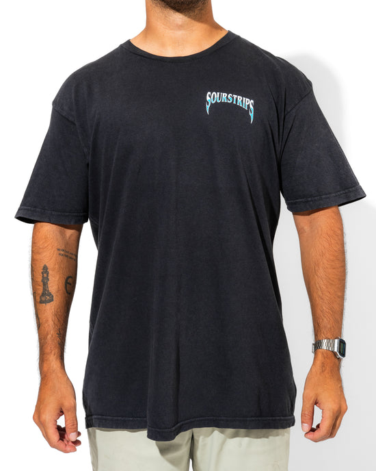 A man wearing the SourStrips Blue Venom Tee—a loose-fitting unisex cotton shirt with "SOUR STRIPS" on the chest—light-colored pants, a wristwatch, and visible arm tattoos stands against a white background.