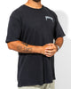A person wearing a loose unisex Blue Venom Tee by SourStrips with "SOURSTRIPS" text on the chest, light pants, a wristwatch, and visible arm tattoos stands confidently against a white background.