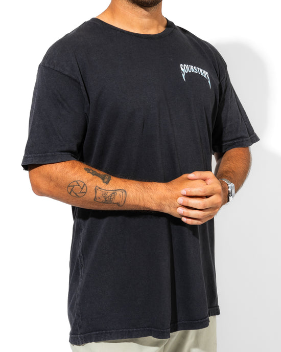 A person wearing a loose unisex Blue Venom Tee by SourStrips with "SOURSTRIPS" text on the chest, light pants, a wristwatch, and visible arm tattoos stands confidently against a white background.