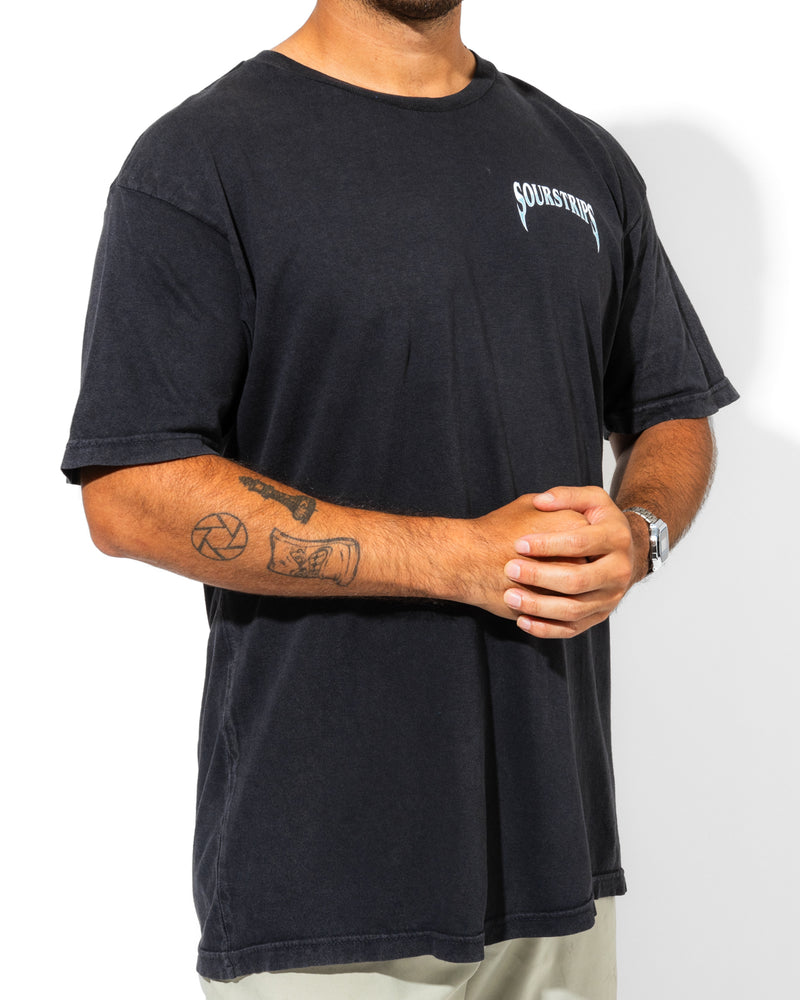 A person wearing a loose unisex Blue Venom Tee by SourStrips with "SOURSTRIPS" text on the chest, light pants, a wristwatch, and visible arm tattoos stands confidently against a white background.