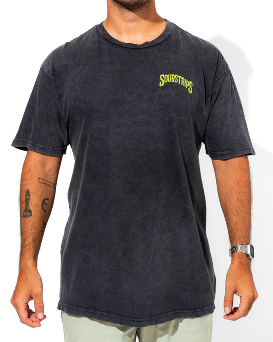 A person wearing a faded black SourStrips Twisted Temptation Tee with yellow-green chest text, light pants, a watch, and visible arm tattoos stands against a white background. This unisex cotton tee adds bold style to any outfit.