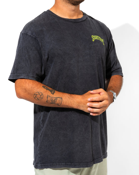 A person stands against a plain white background, wearing the SourStrips Twisted Temptation Tee—a faded black unisex cotton shirt with yellow chest text—hands clasped, tattoos on their right forearm, and a watch on their left wrist.