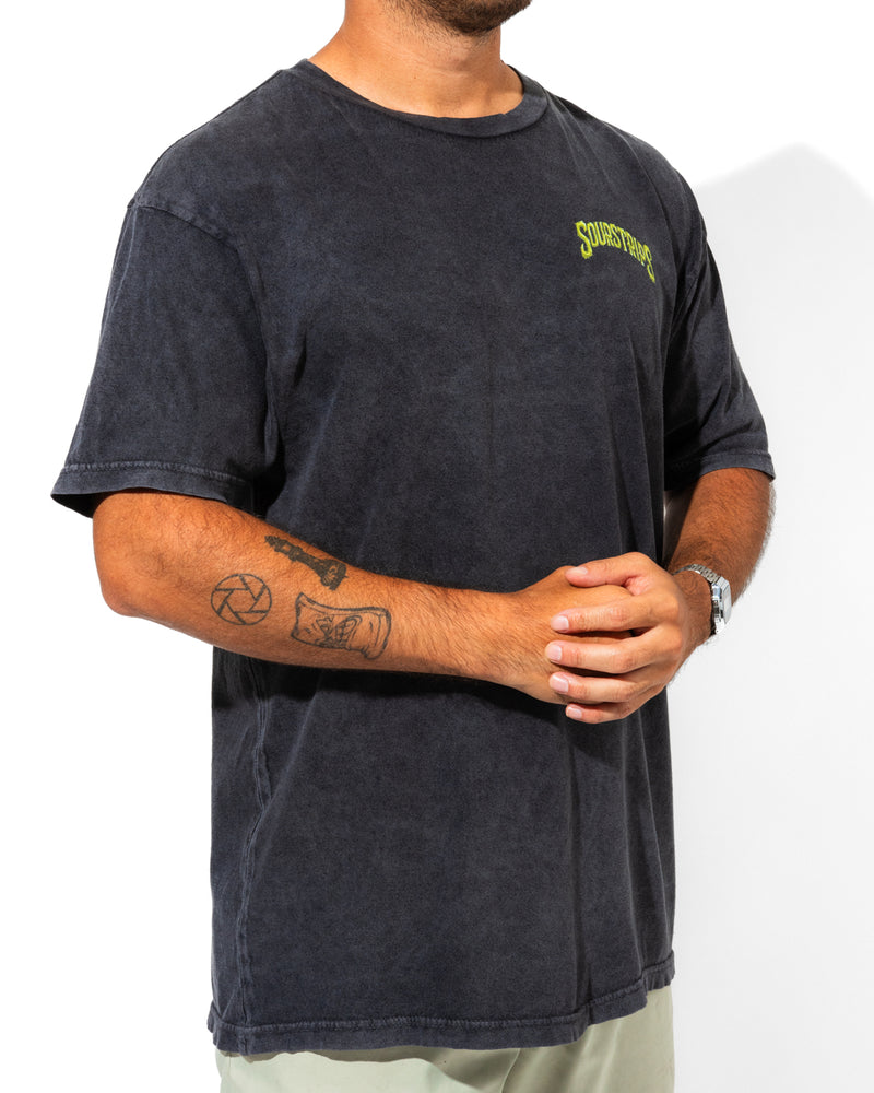 A person stands against a plain white background, wearing the SourStrips Twisted Temptation Tee—a faded black unisex cotton shirt with yellow chest text—hands clasped, tattoos on their right forearm, and a watch on their left wrist.