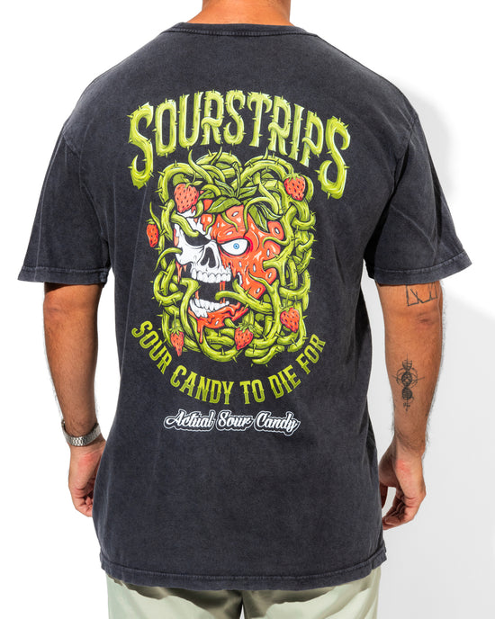 A person wearing the SourStrips Twisted Temptation Tee, a black unisex cotton shirt with a colorful skull, strawberries, and green vines on the back, and text reading "Sourstrips Sour Candy to Die For" and "Actual Sour Candy.