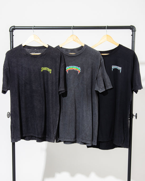 Three black unisex cotton Twisted Temptation Tees by SourStrips hang on wooden hangers from a metal rack. Each sports a vibrant "Sourcefurs" logo on the left chest, set against a plain white background to highlight these graphic T-shirts.
