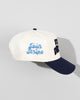The SourStrips Dreamberry Hat-White features a white and navy design with a blue embroidered side logo, partially visible dark blue front lettering, a curved brim, and ventilation eyelets for added comfort.