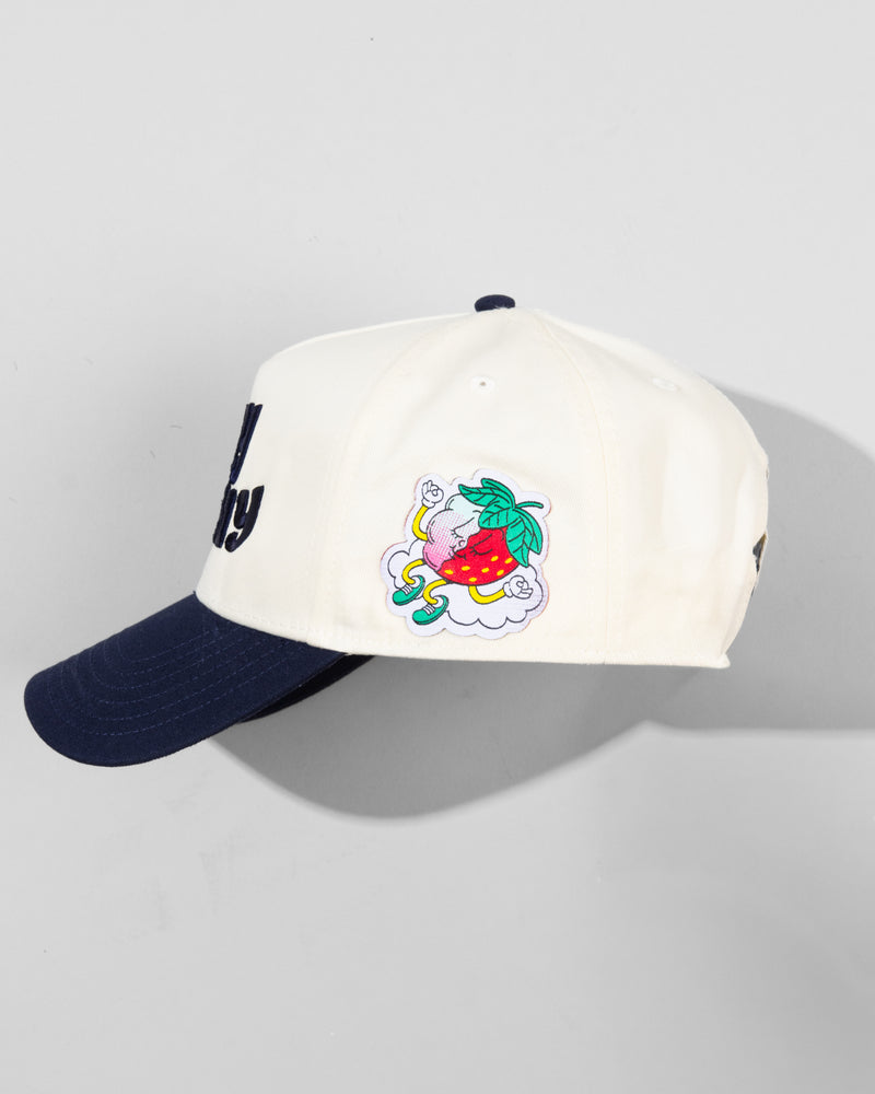 The SourStrips Dreamberry Hat-White features a cream crown, navy brim, colorful embroidered gray cat on a red strawberry with green leaves, and a yellow-detailed logo on the side.