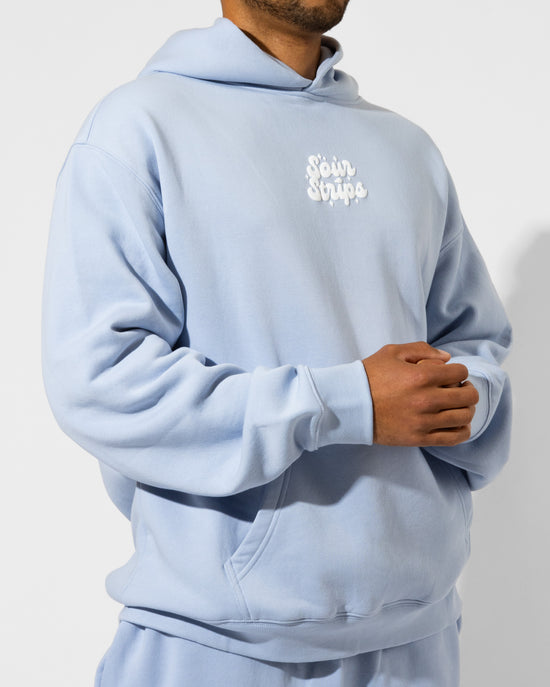 A person in a light blue unisex Dreamberry Hoodie by SourStrips with a white embroidered logo on the chest stands with their hands clasped against a plain white background.