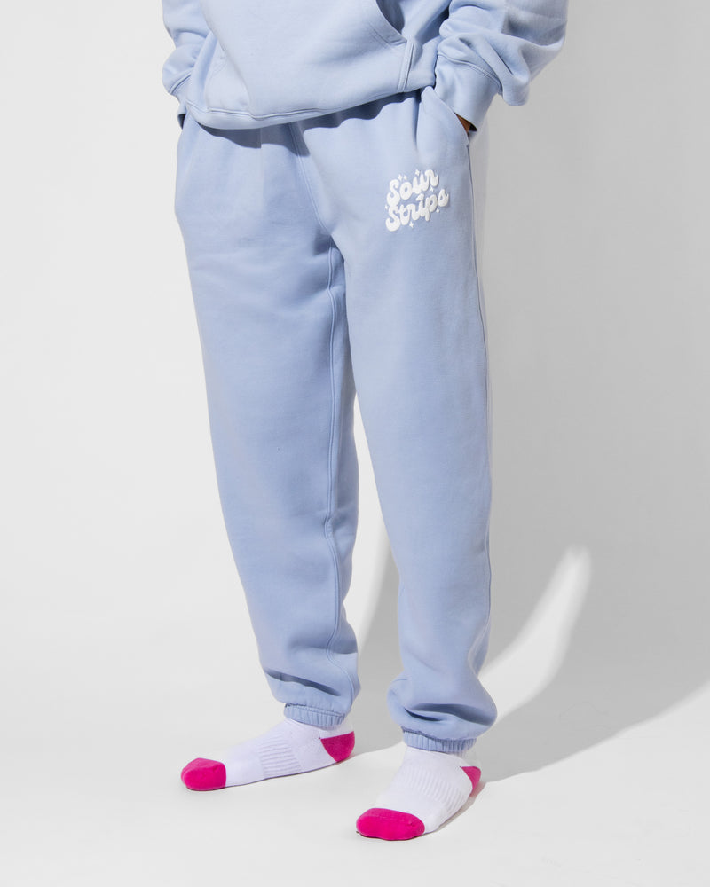 A person is shown from the waist down wearing SourStrips Dreamberry Joggers in light blue with the "Sour Strips" logo and a matching sweatshirt, paired with white socks featuring pink toes and heels. Hands are in pockets.