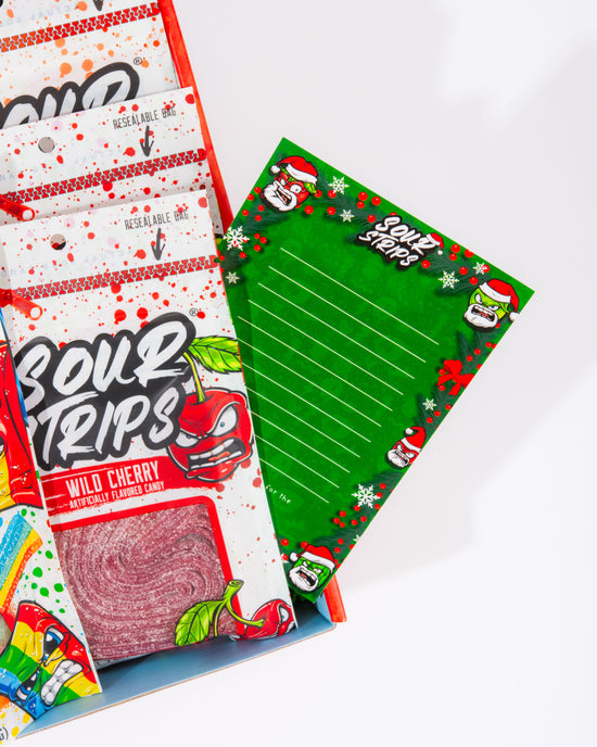 Close up of the blank green sour strips card with holiday characters on it. It is sticking out of the holiday box and you can see a bag of cherry sour strips laying in the box.