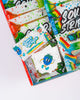 Close up of the sticker pack and some sour strips inside the box. You can see the retro blue raspberry character on the sticker pack and it is laying on a bag of watermelon sour strips.