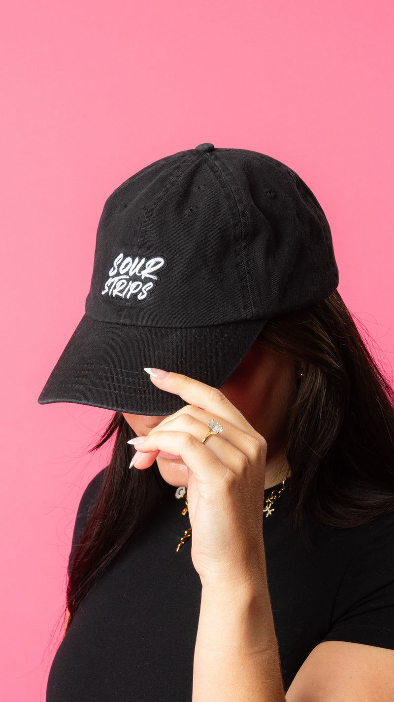 Girl wearing hat looking down with hand on brim wearing a ring. Black hat with curved brim. Sour Strips logo on front center of hat in white color. Girl is wearing gold necklace and has medium to long length dark hair.