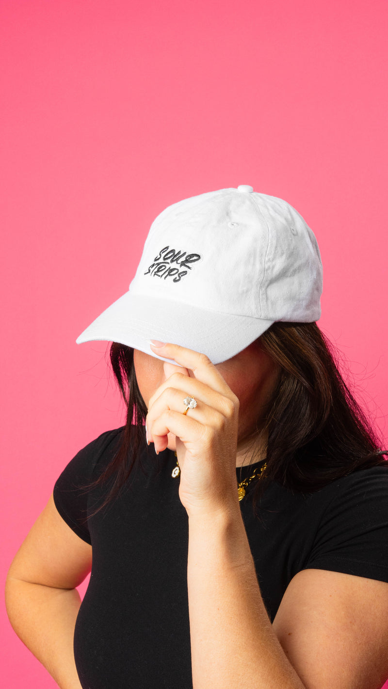 girl wearing white hat with curved brim. She has her hand on the brim of the hat wearing a ring. Sour Strips logo on front center of hat in white black color. Girl has medium to long dark hair and is wearing a gold necklace and black tshirt.