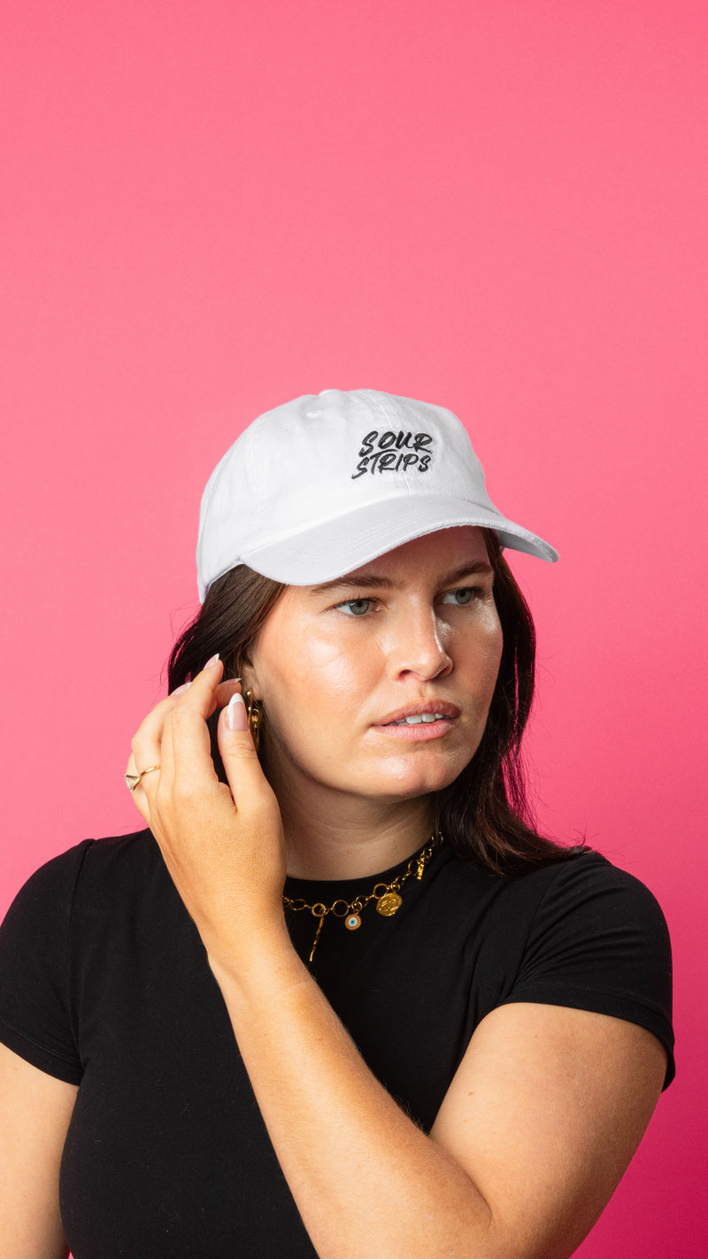 girl wearing white hat with curved brim. She is looking away from camera and is brushing back her hair. Sour Strips logo on front center of hat in white black color. Girl has medium to long dark hair and is wearing a gold necklace and black tshirt.