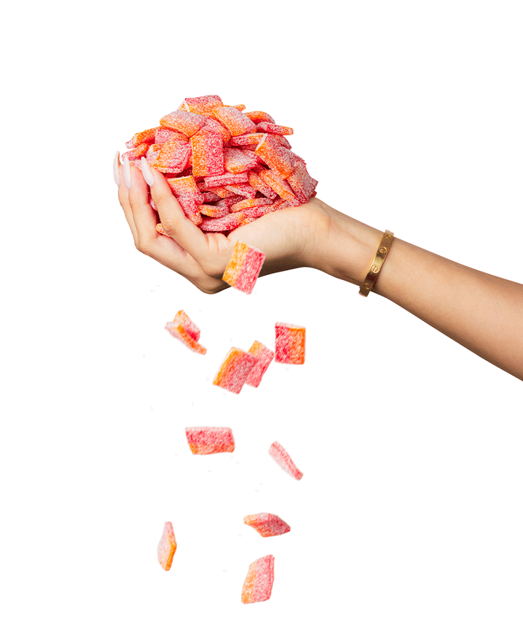 A hand, adorned with a bracelet, holds a pile of red and orange sugar-coated gummy candies from the Sour Strips Bites-Four Pack by Sour Strips, with some candies falling down from the pile. The background is white.