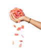 A hand, adorned with a bracelet, holds a pile of red and orange sugar-coated gummy candies from the Sour Strips Bites-Four Pack by Sour Strips, with some candies falling down from the pile. The background is white.