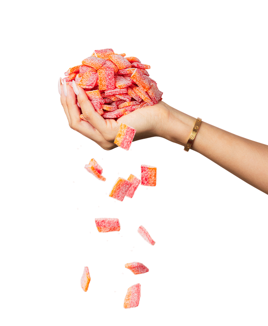 A hand, adorned with a bracelet, holds a pile of red and orange sugar-coated gummy candies from the Sour Strips Bites-Four Pack by Sour Strips, with some candies falling down from the pile. The background is white.