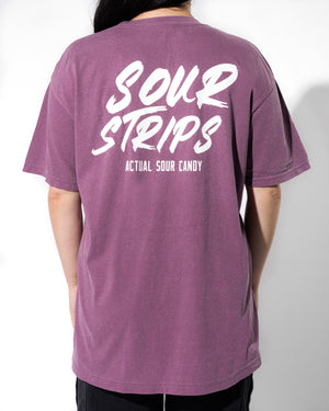 A person is shown from behind wearing an oversized, box-fit Logo Tee - Berry by SourStrips. The T-shirt features "SOUR STRIPS" in large white letters and "ACTUAL SOUR CANDY" in smaller print beneath it. The person has long, dark hair and is standing against a plain, light-colored background.