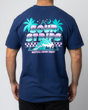A person is wearing the Night Sky Tee, a dark blue unisex t-shirt by SourStrips, featuring a vibrant graphic on the back. The design showcases palm trees, a wave, and retro-style text that reads "Sour Strips" above and "Actual Sour Candy" below. The person has a tattoo on their right forearm.
