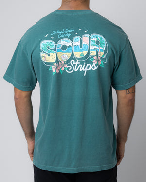 A person is wearing the SourStrips Greetings Tee, an oversized fit unisex T-shirt in a teal color, featuring a colorful design on the back. The design displays text that reads "Actual Sour Candy: Sour Strips" surrounded by tropical-themed illustrations including flowers, palm trees, and a sun with birds.