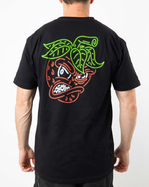 A person is wearing the SourStrips brand Neon Tee - Strawberry, a black, relaxed fit T-shirt featuring a colorful, neon-style graphic of an angry-faced strawberry with green leaves, red fruit, and visible teeth on the back. The Cotton Unisex design stands out against the plain white background.