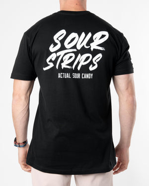 A person is seen wearing a SourStrips "Logo Tee - Black" featuring white text on the back that reads "Sour Strips" in large font and "Actual Sour Candy" in smaller font below it. The individual is facing away from the camera, showcasing the oversized box fit of their graphic T-shirt, revealing only their upper body.